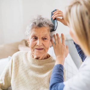 An unrecognizable health visitor combing hair of senior woman sitting on a sofa at home.